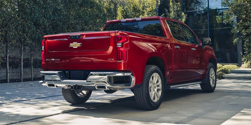 A red Chevy Silverado viewed from the trunk side. It is parked in an urban parking lot in front of an office building.